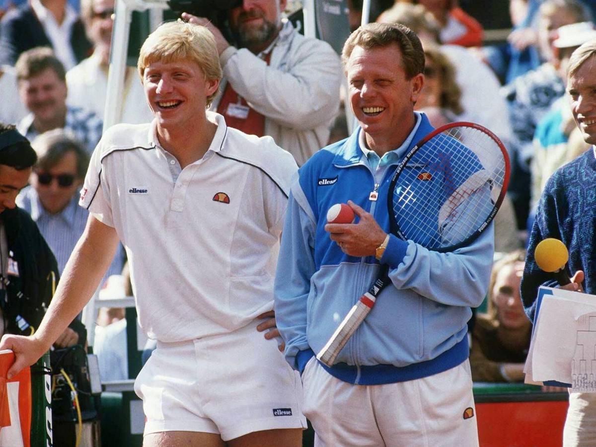 Boris Becker und Günther Bosch stehen auf dem Tennisplatz. Becker lehnt sich auf das Netz, Bosch hält einen Schläger und einen Ball.