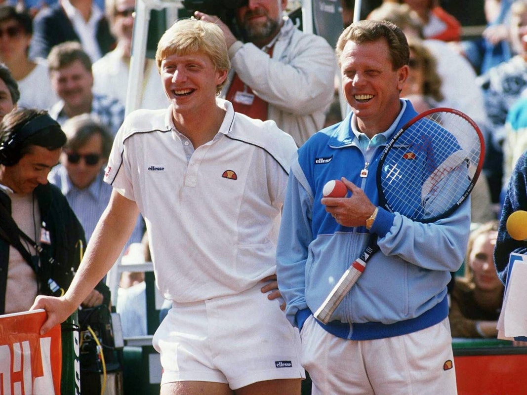 Boris Becker und Günther Bosch stehen auf dem Tennisplatz. Becker lehnt sich auf das Netz, Bosch hält einen Schläger und einen Ball.
