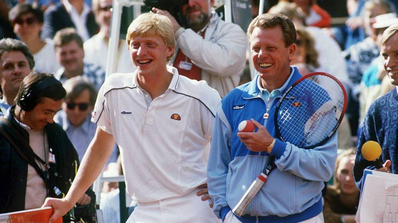 Boris Becker und Günther Bosch stehen auf dem Tennisplatz. Becker lehnt sich auf das Netz, Bosch hält einen Schläger und einen Ball.