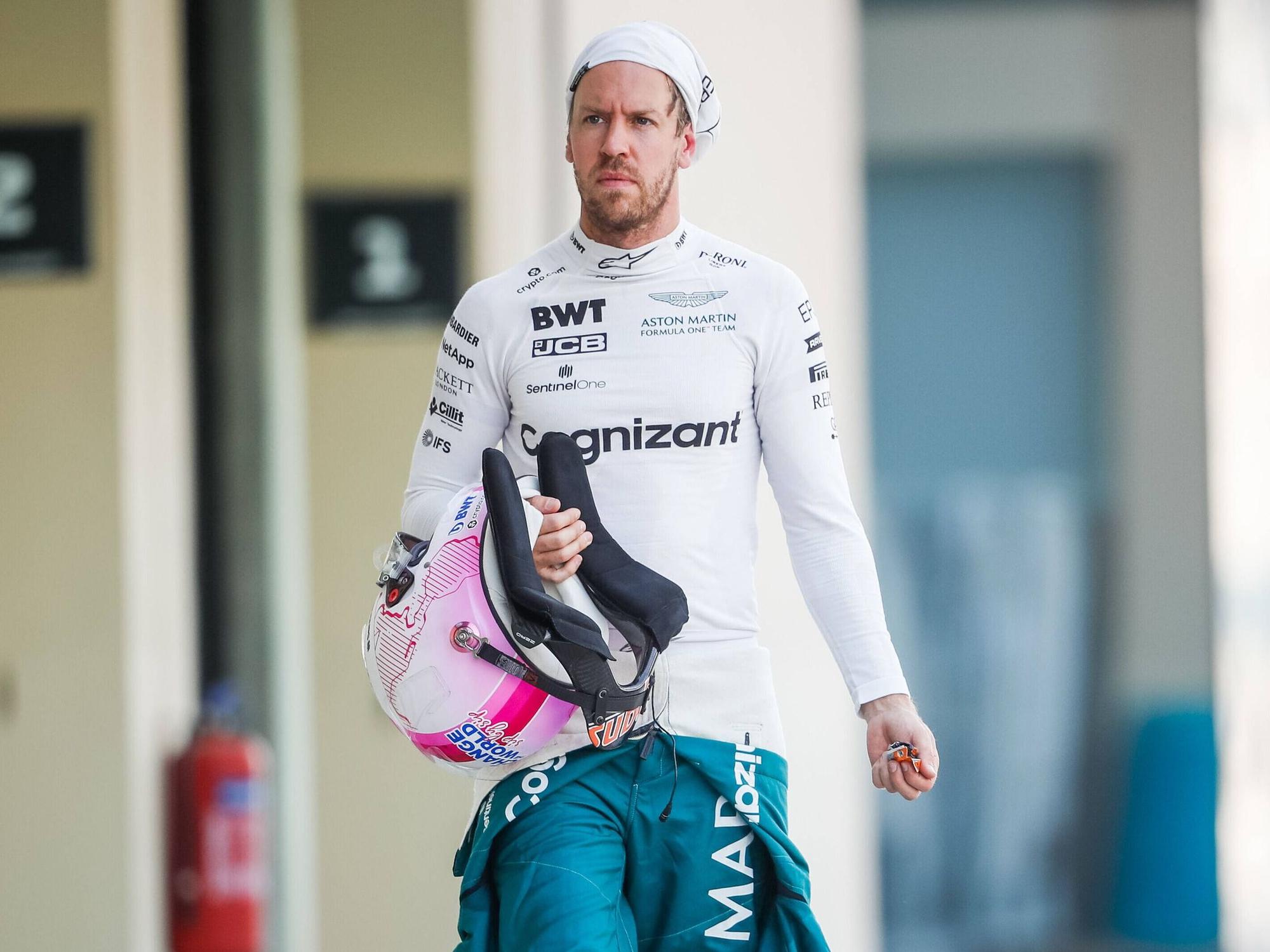 Sebastian Vettel hat seinen Schutzanzug halb ausgezogen, in der Hand hält er seinen Helm. Die Feuermaske trägt er hochgezogen.