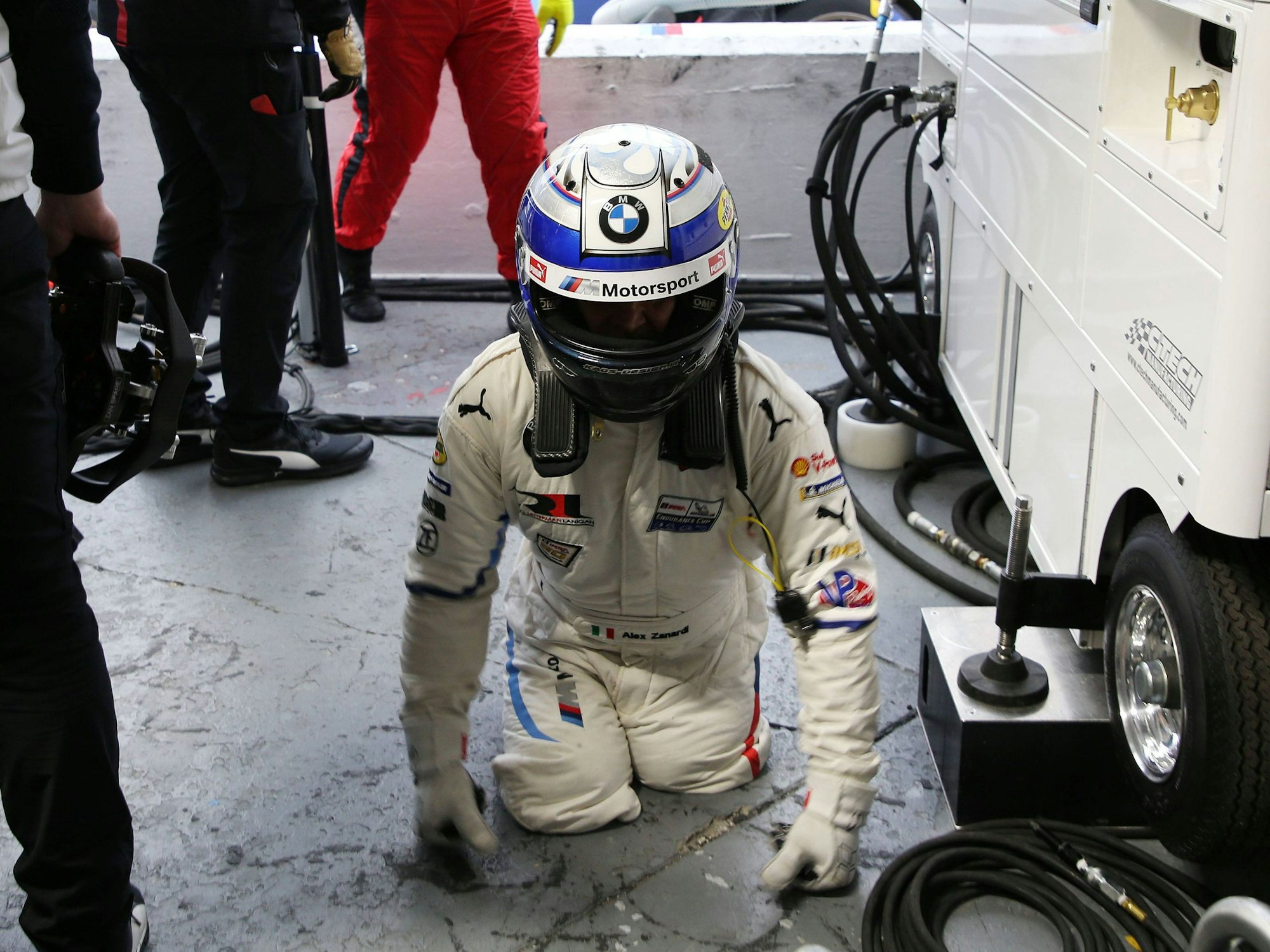 Alessandro Zanardi ist in Rennfahrermontur in der Box. Er steht auf seinen Oberschenkeln und stützt sich mit seinen Händen ab.