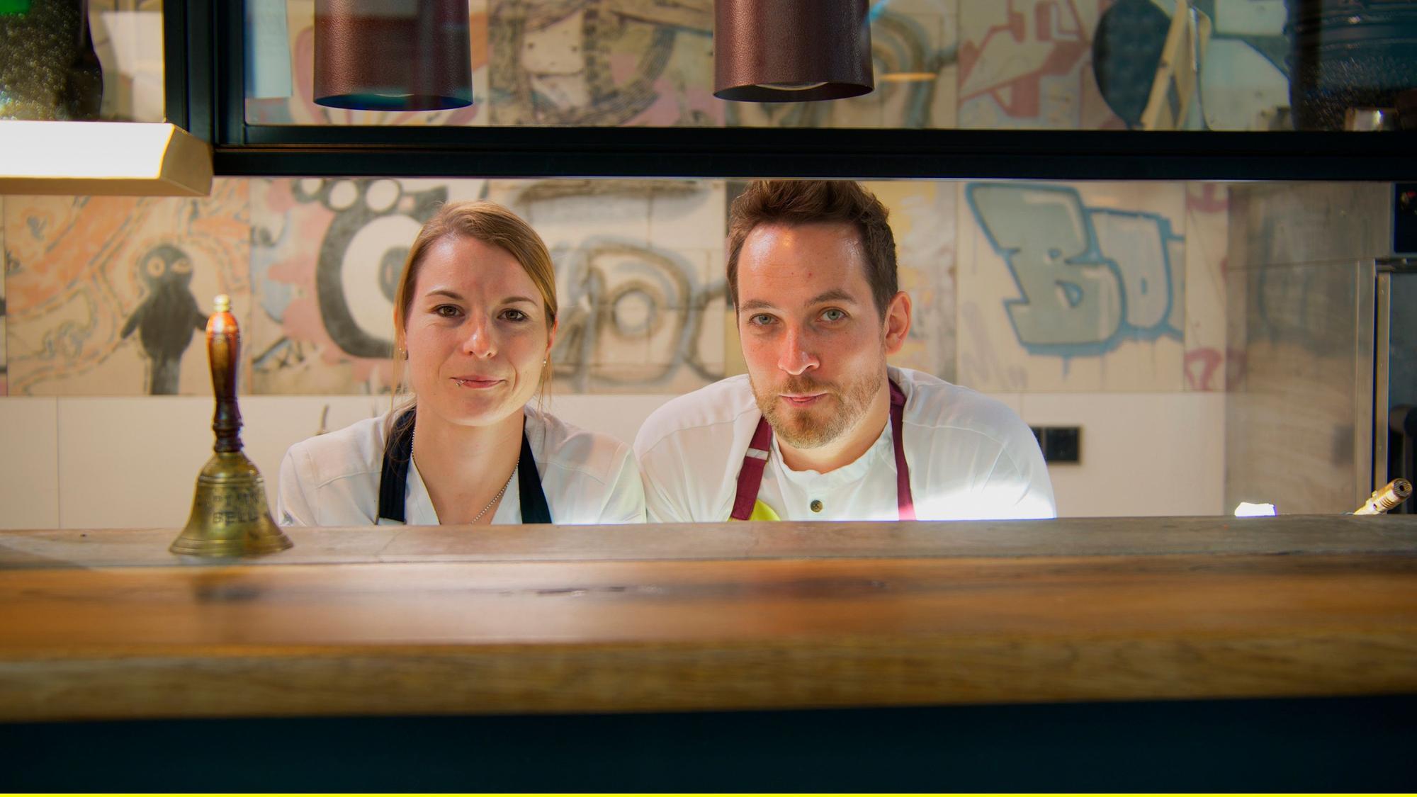 Sonja Baumann und Erik Scheffler schauen durch die Durchreiche aus der Küche.