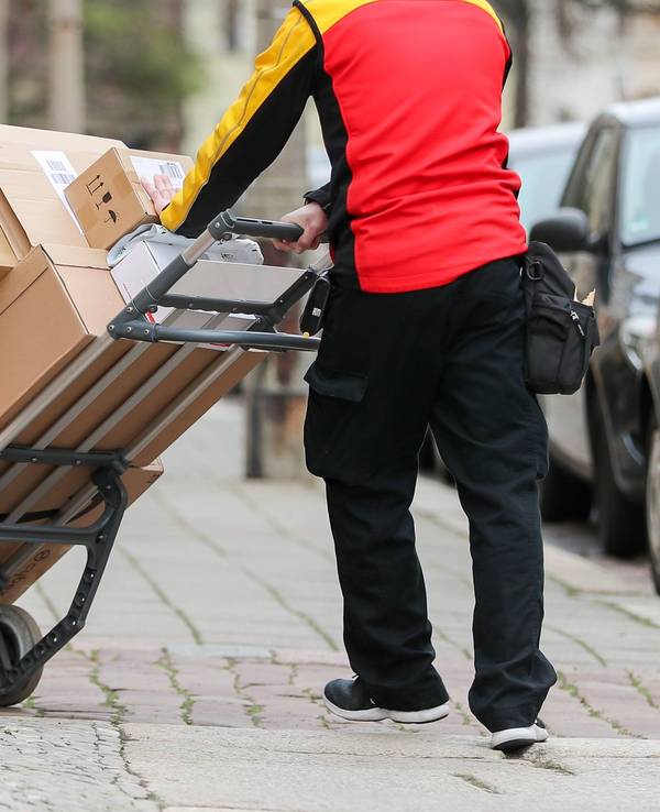 Ein Paketzusteller geht mit einer Sackkarre voller Pakete zu einem Haus.