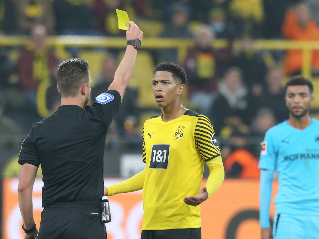 Jude Bellingham bekommt vom Schiedsrichter die Gelbe Karte. Bellingham schaut verständnislos und breitet die Arme fragend aus.