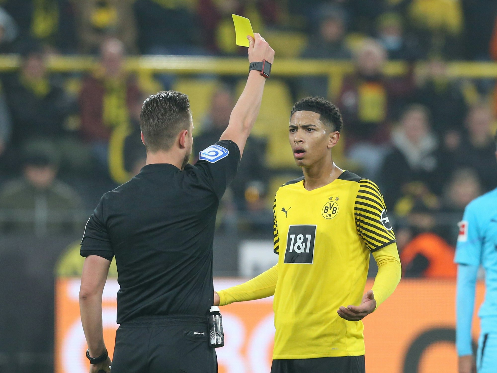 Jude Bellingham bekommt vom Schiedsrichter die Gelbe Karte. Bellingham schaut verständnislos und breitet die Arme fragend aus.