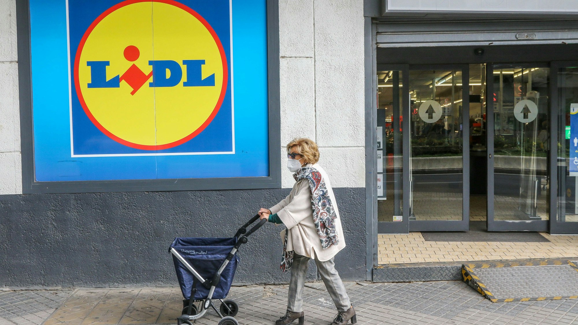 Eine Frau mit Rollator trägt einen Mund-Nasen-Schutz und passiert eine Lidl-Filiale.
