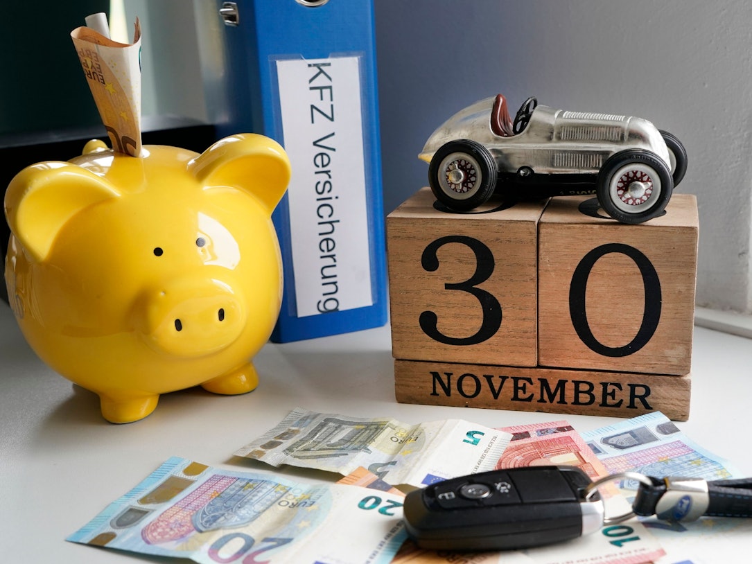 Auf dem Foto sieht man mehrere Geldscheine, ein gelbes Sparschwein, ein Autoschlüssel und einen Aktenordner.