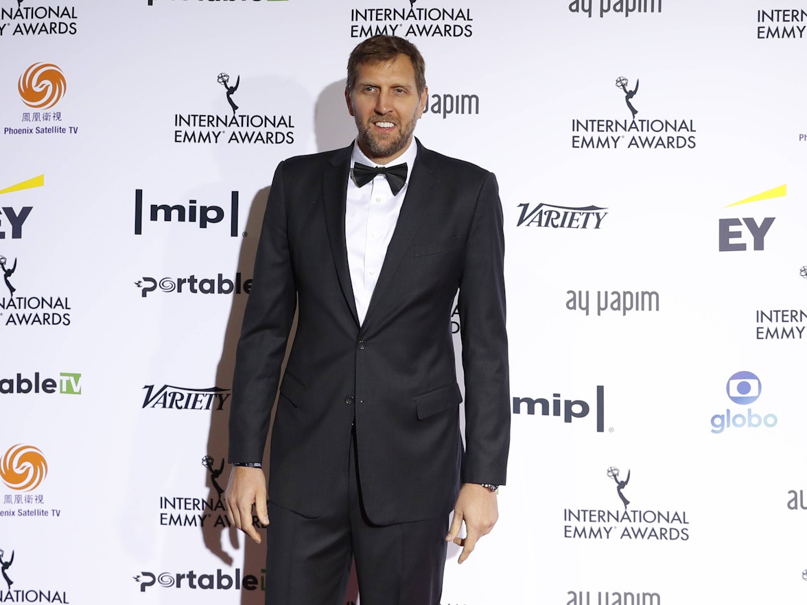 Dirk Nowitzki steht im Anzug vor einer Werbebande.