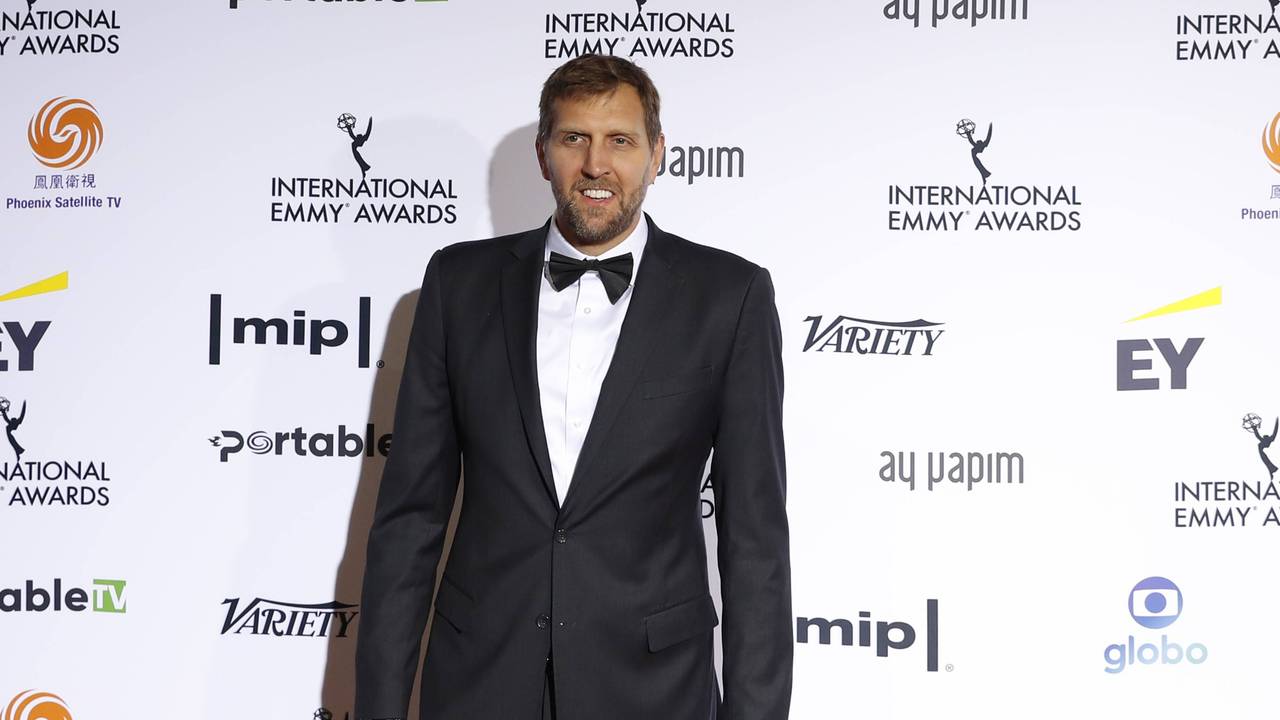 Dirk Nowitzki steht im Anzug vor einer Werbebande.