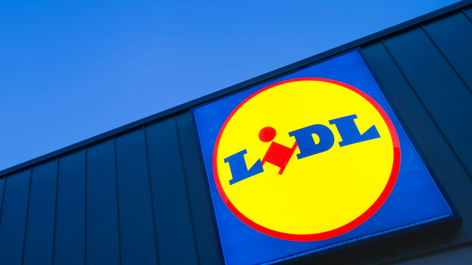 Das Logo eines Lidl-Supermarktes in Berlin-Reinickendorf leuchtet vor dem Abendhimmel.