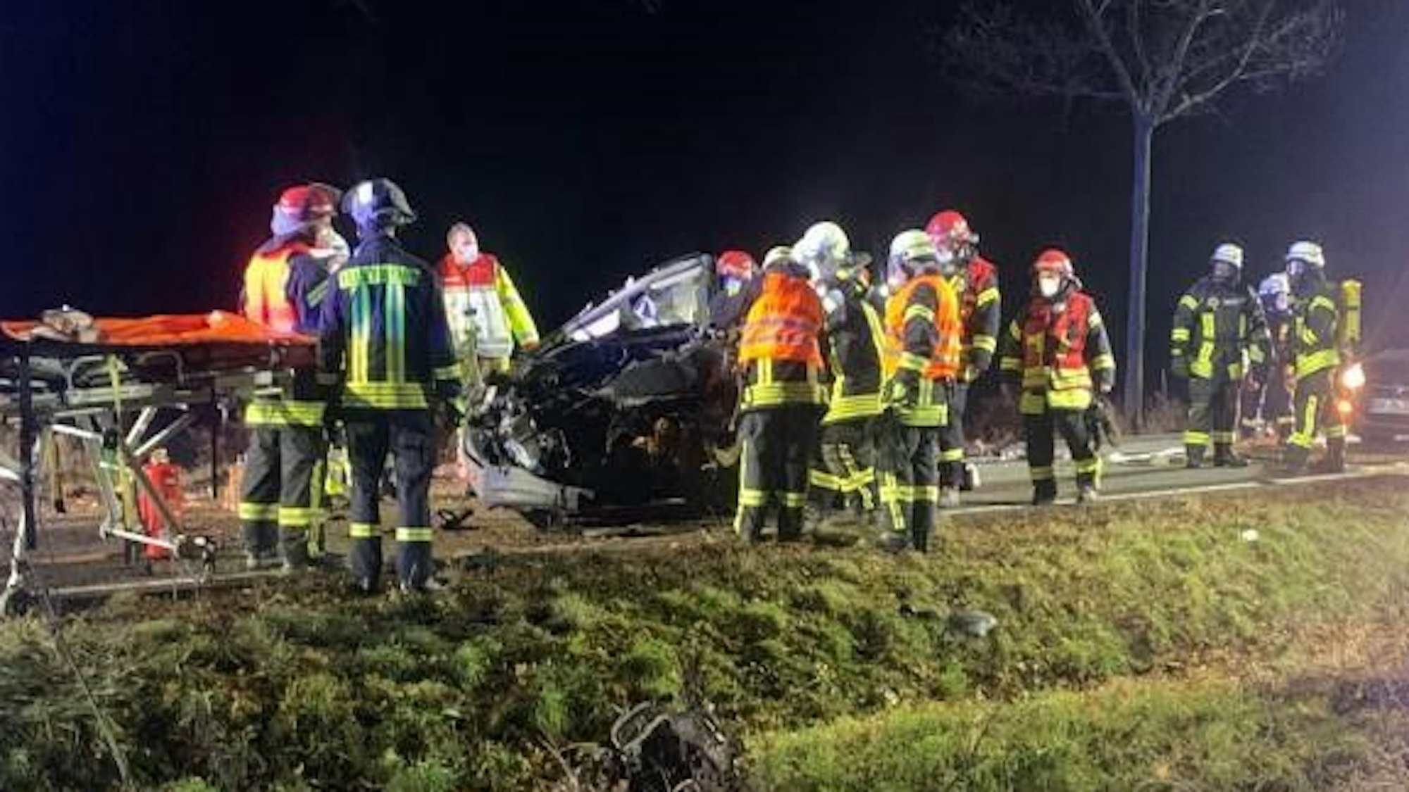 Einsatzkräfte der Feuerwehr stehen am Wrack auf der Straße.