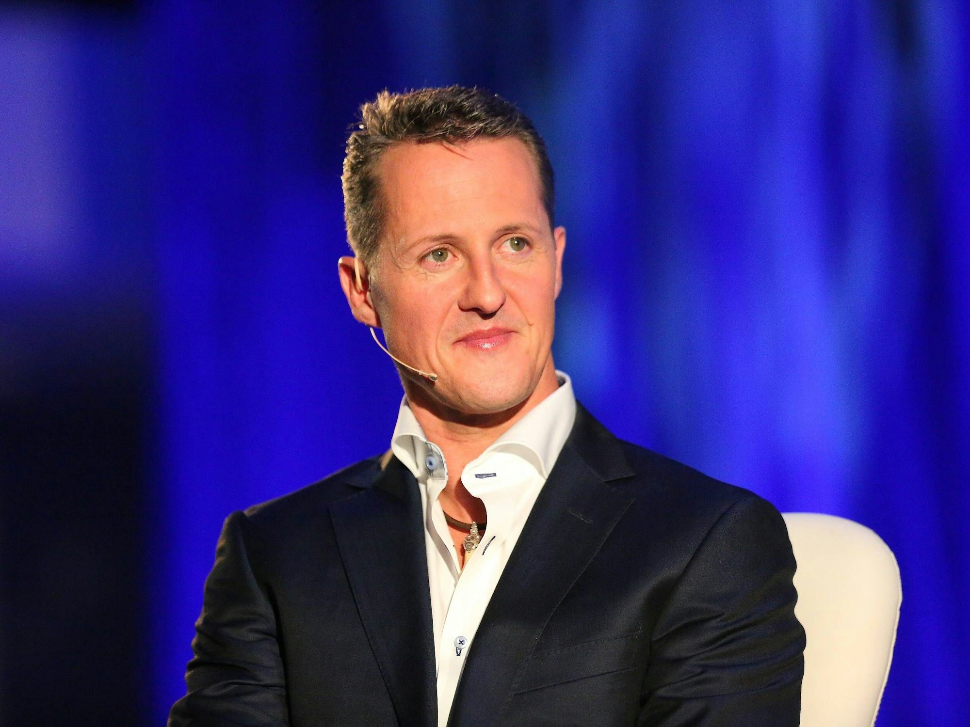 Michael Schumacher sitzt auf einem Stuhl. Er trägt Hemd und Anzug.