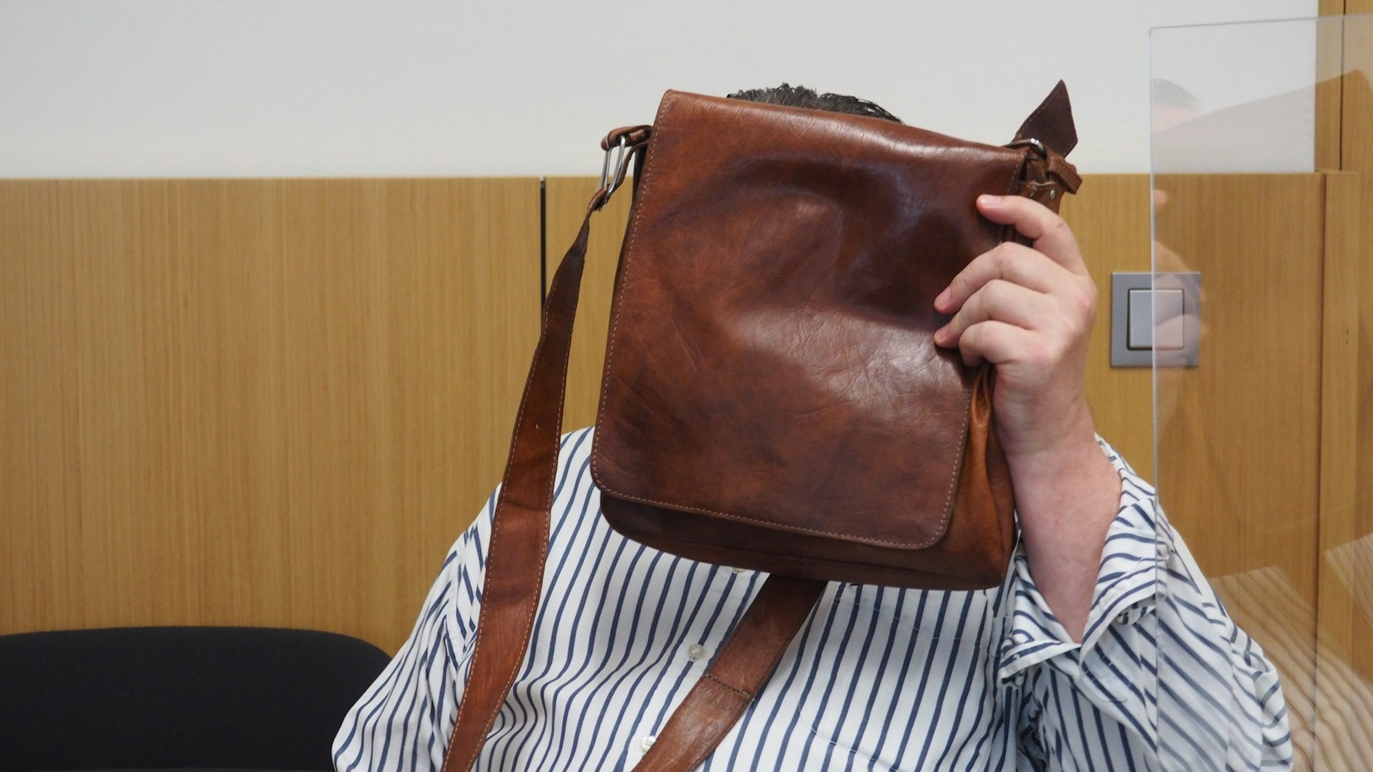 Hinter einer braunen Herren-Schultertasche verbirgt der angeklagte Betrüger im Düsseldorfer Gerichtssaal sein Gesicht.