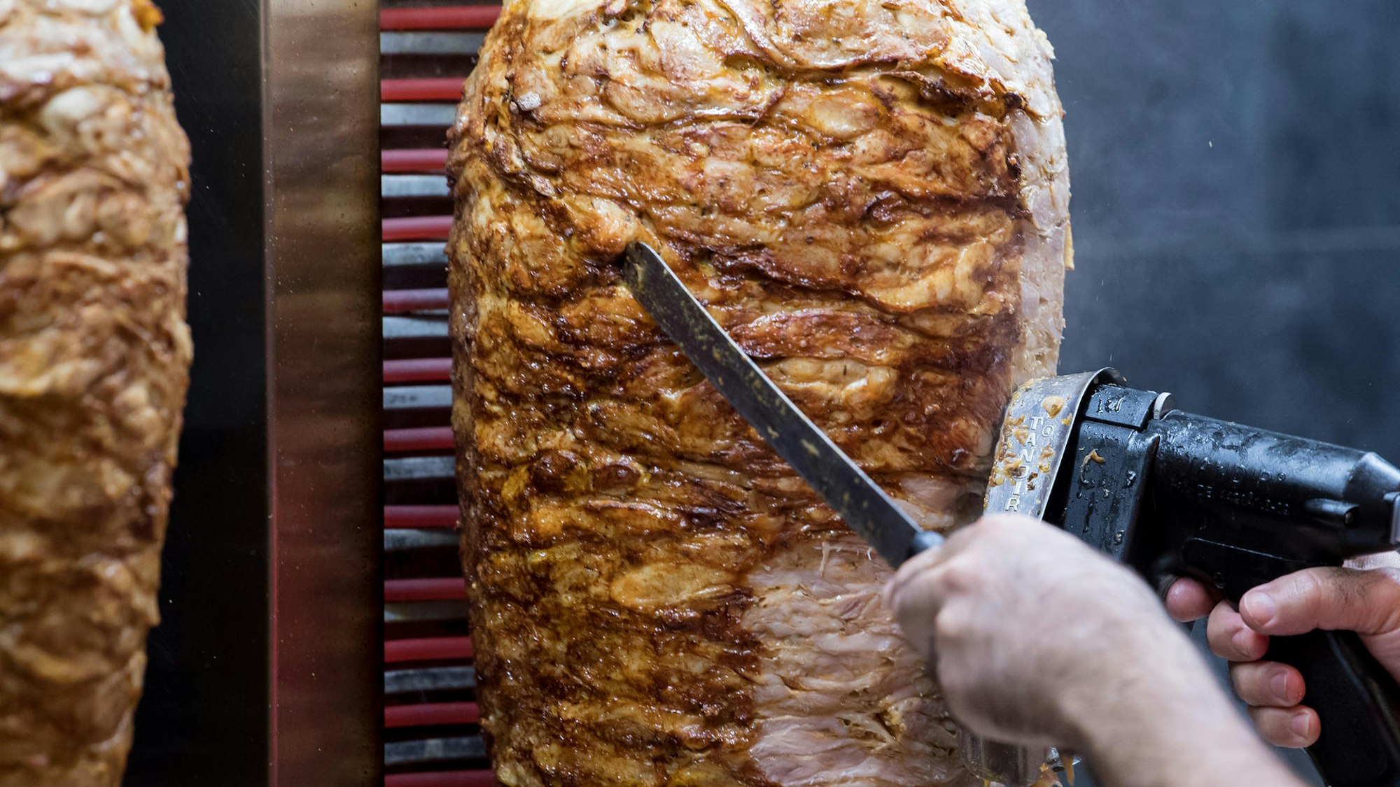 Dönerspieße drehen sich in einem Imbiss, ein Mann schneidet Stücke von einem der Spieße herunter. Das Lieblingsessen der Deutschen (auf dem Foto von einem Münchner Imbiss) – ist heute ein Milliardengeschäft, ein Großteil aller Spieße wird industriell hergestellt.