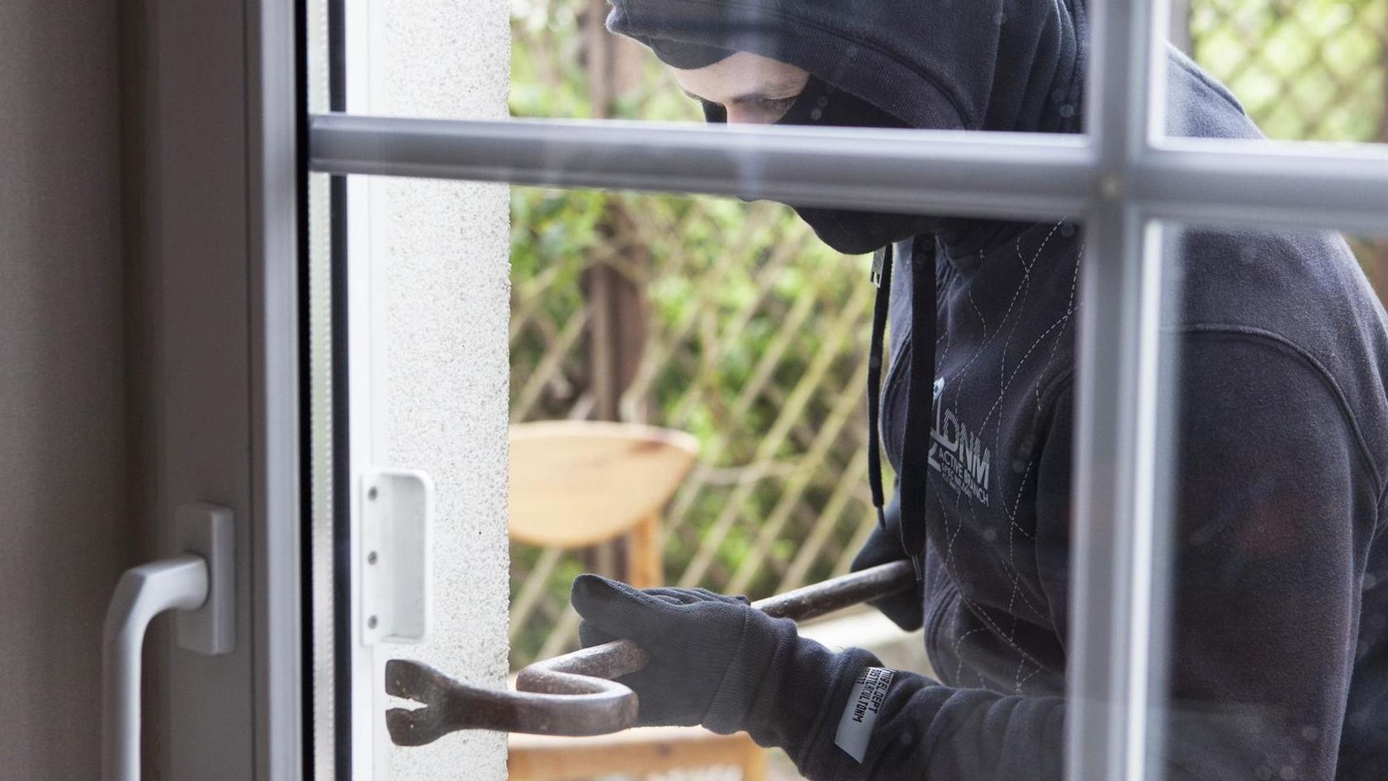 Ein maskierter Einbrecher setzt an einer Glastür von außen eine Brechstange an.