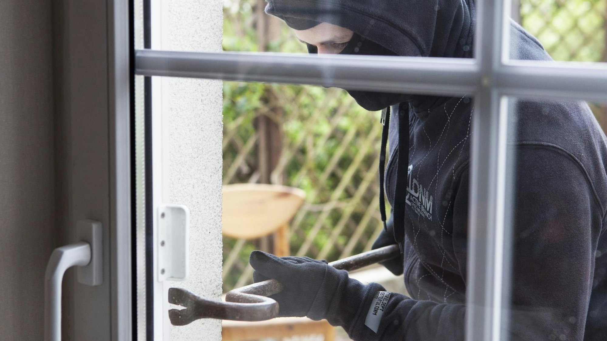 Ein maskierter Einbrecher setzt an einer Glastür von außen eine Brechstange an.