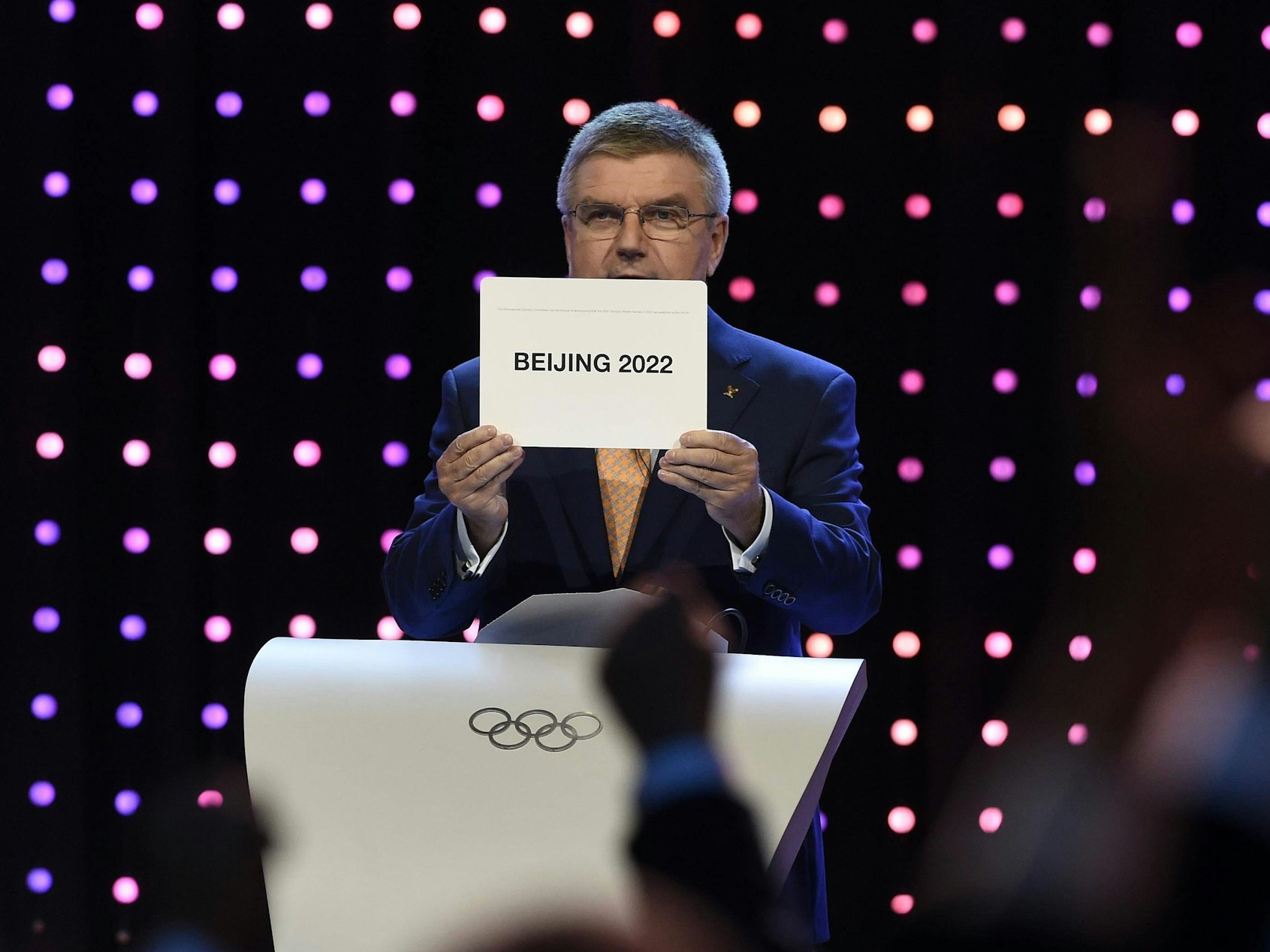 Thomas Bach hält ein Schild hoch, auf dem „Beijing 2022“ steht.