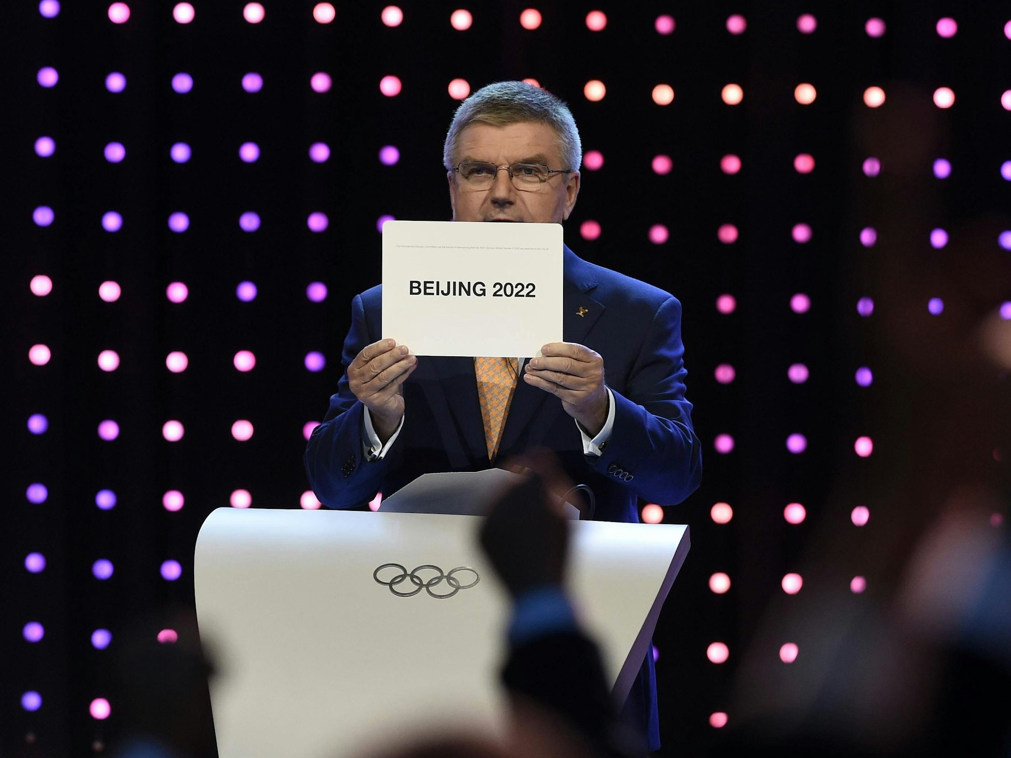 Thomas Bach hält ein Schild hoch, auf dem „Beijing 2022“ steht.