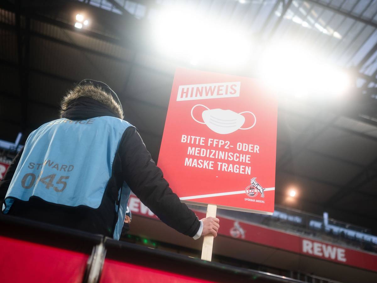 Ein Ordner steht auf der Tribüne und hält ein Schild, das auf die Pflicht zum Tragen eines Mund-Nasen-Schutzes hinweist.
