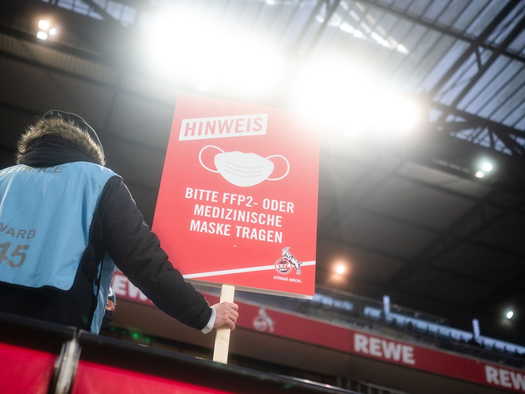 Ein Ordner steht auf der Tribüne und hält ein Schild, das auf die Pflicht zum Tragen eines Mund-Nasen-Schutzes hinweist.