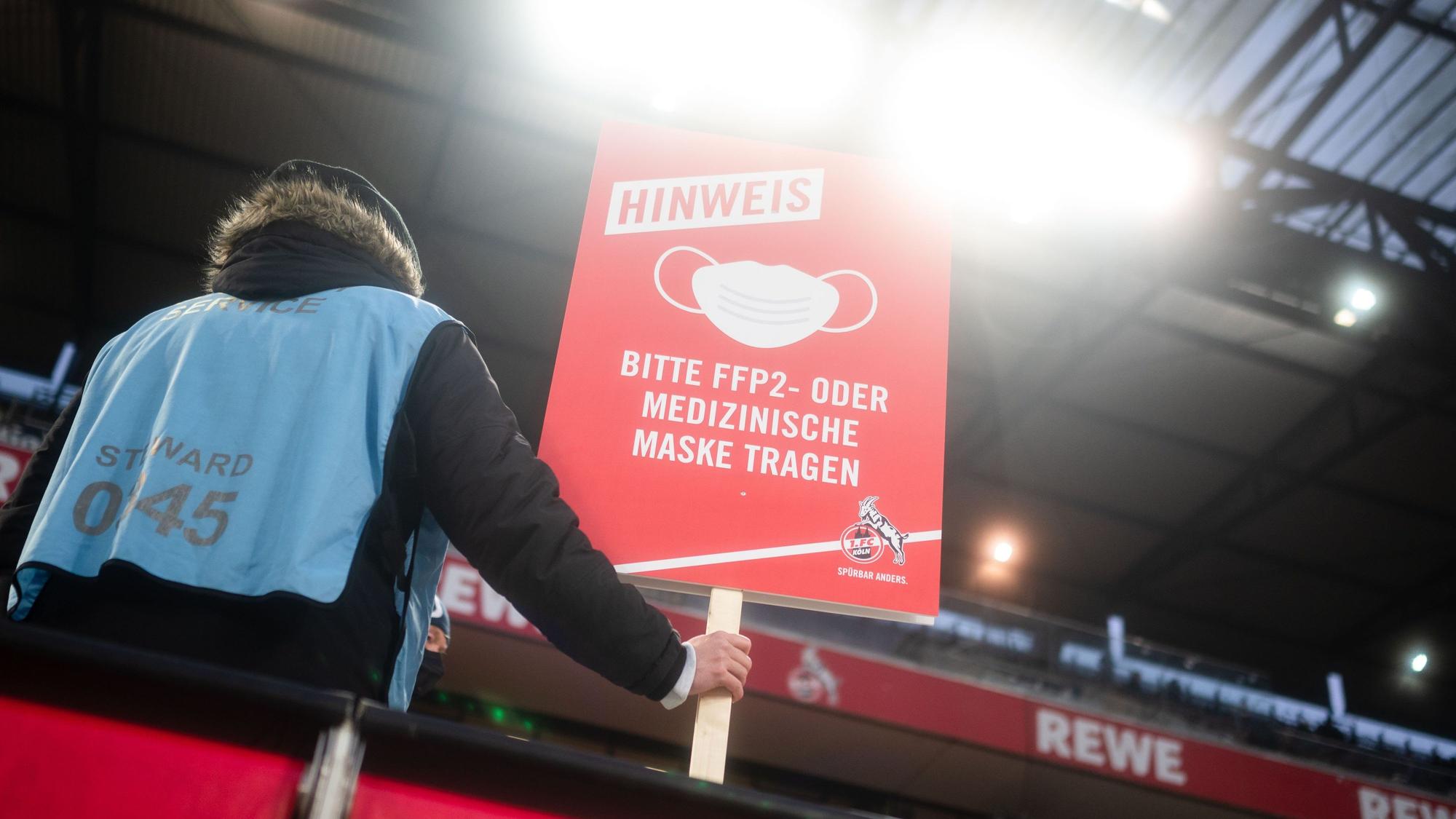 Ein Ordner steht auf der Tribüne und hält ein Schild, das auf die Pflicht zum Tragen eines Mund-Nasen-Schutzes hinweist.