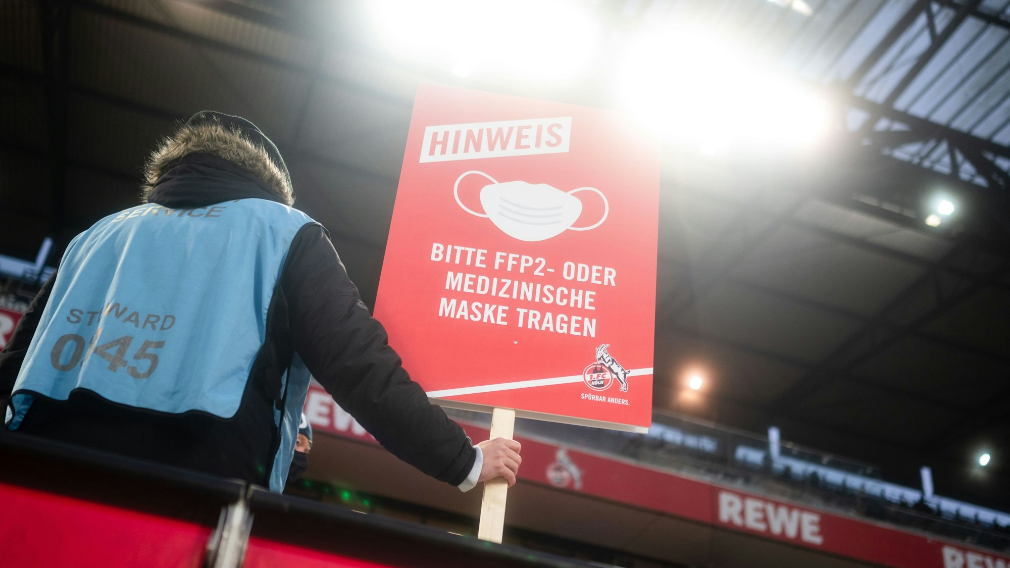 Ein Ordner steht auf der Tribüne und hält ein Schild, das auf die Pflicht zum Tragen eines Mund-Nasen-Schutzes hinweist.