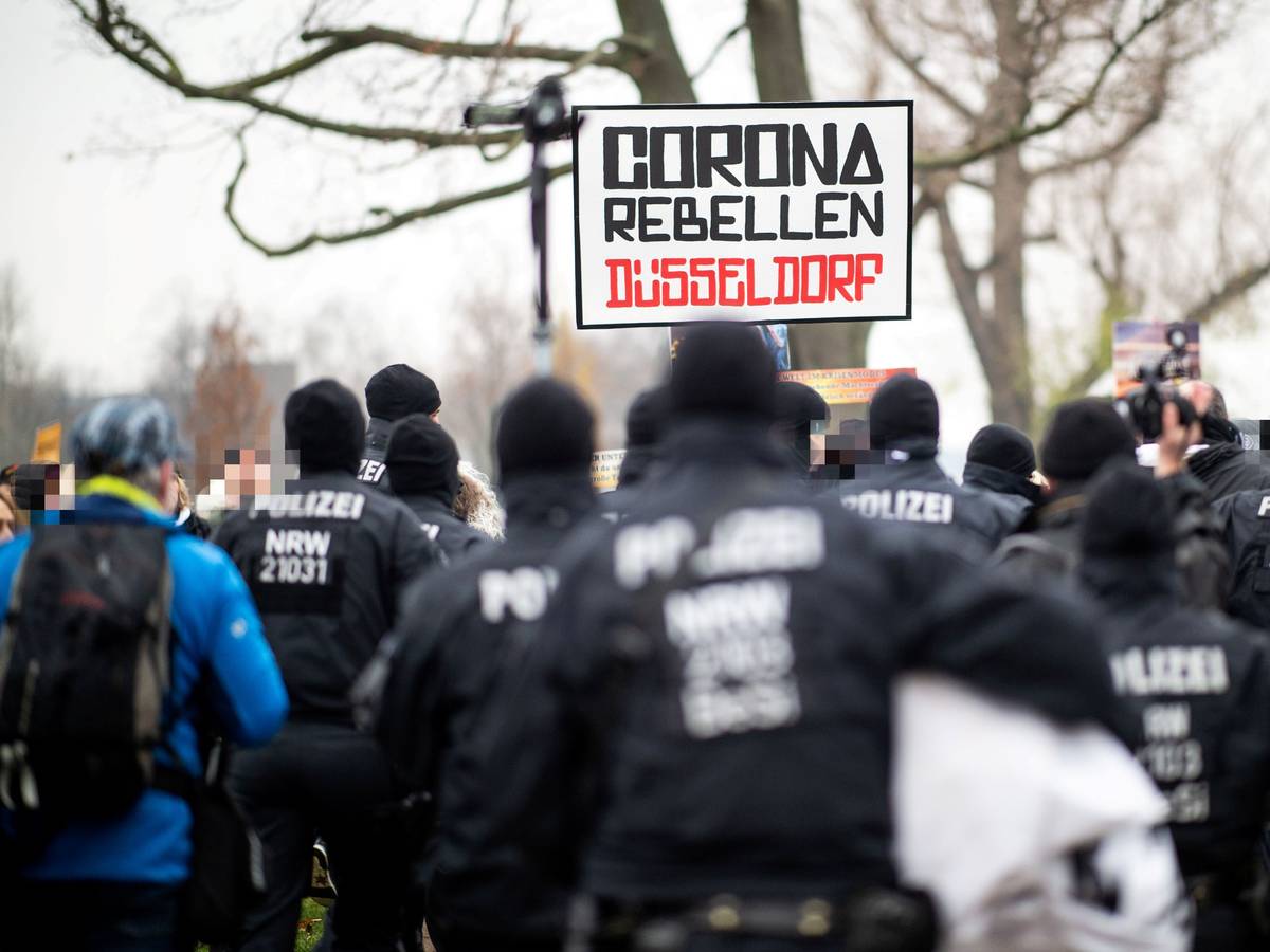 Demonstranten halten bei einer Demonstration am Rheinufer ein Schild mit der Aufschrift „Corona Rebellen Düsseldorf“ in die Höhe, davor stehen Polzisten. D