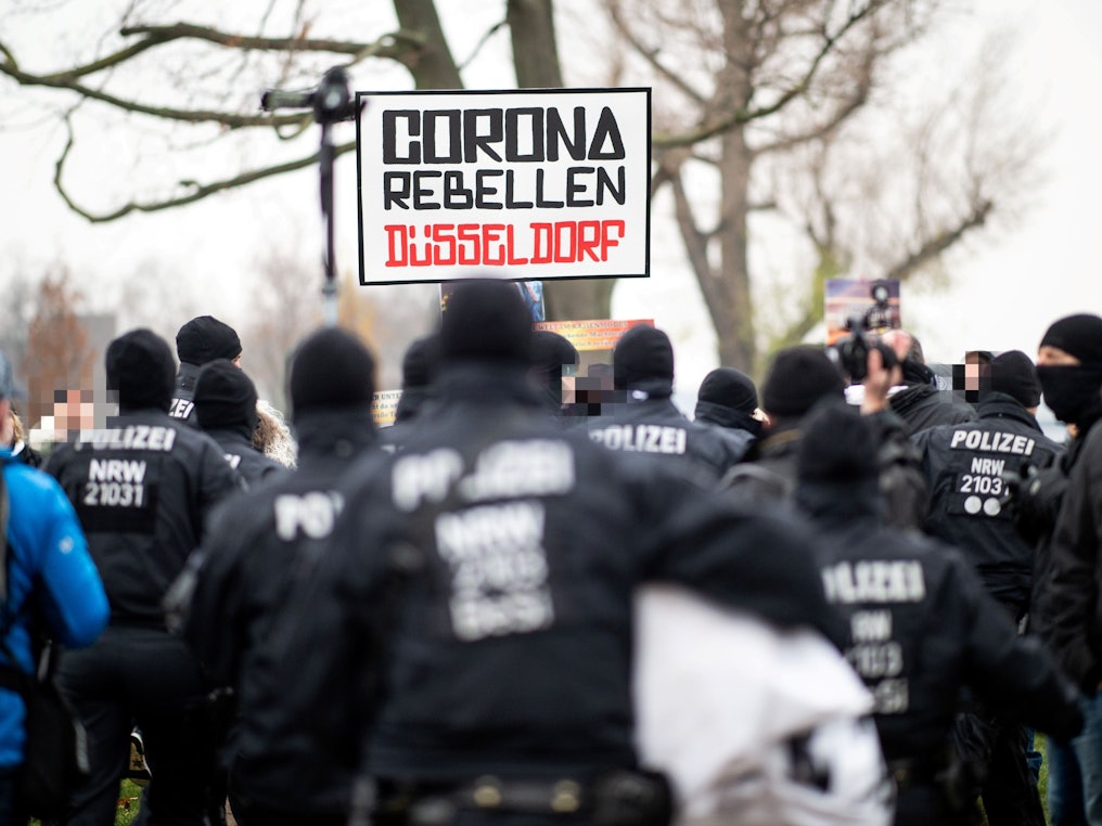 Demonstranten halten bei einer Demonstration am Rheinufer ein Schild mit der Aufschrift „Corona Rebellen Düsseldorf“ in die Höhe, davor stehen Polzisten. D