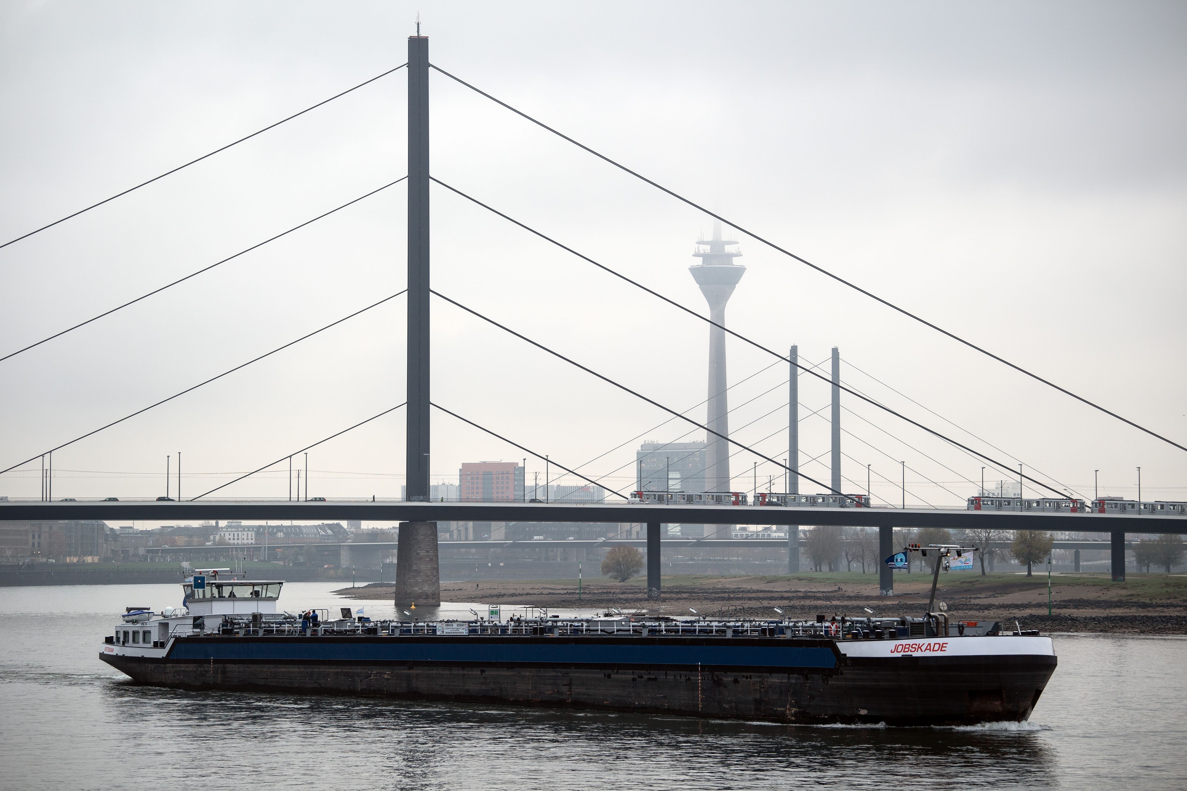 Ein Frachtschiff fährt auf dem Rhein vor der Kulisse der Rheinbrücken und des Rheinturms, der im Dunst beinahe verschwindet.