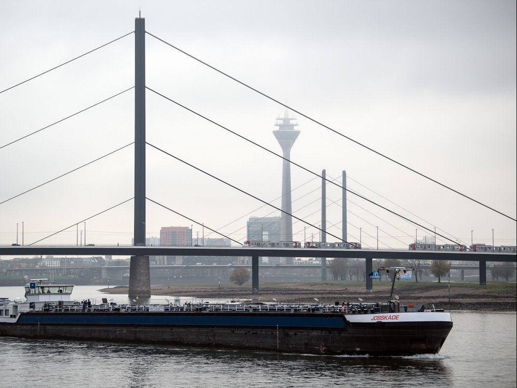 Ein Frachtschiff fährt auf dem Rhein vor der Kulisse der Rheinbrücken und des Rheinturms, der im Dunst beinahe verschwindet.