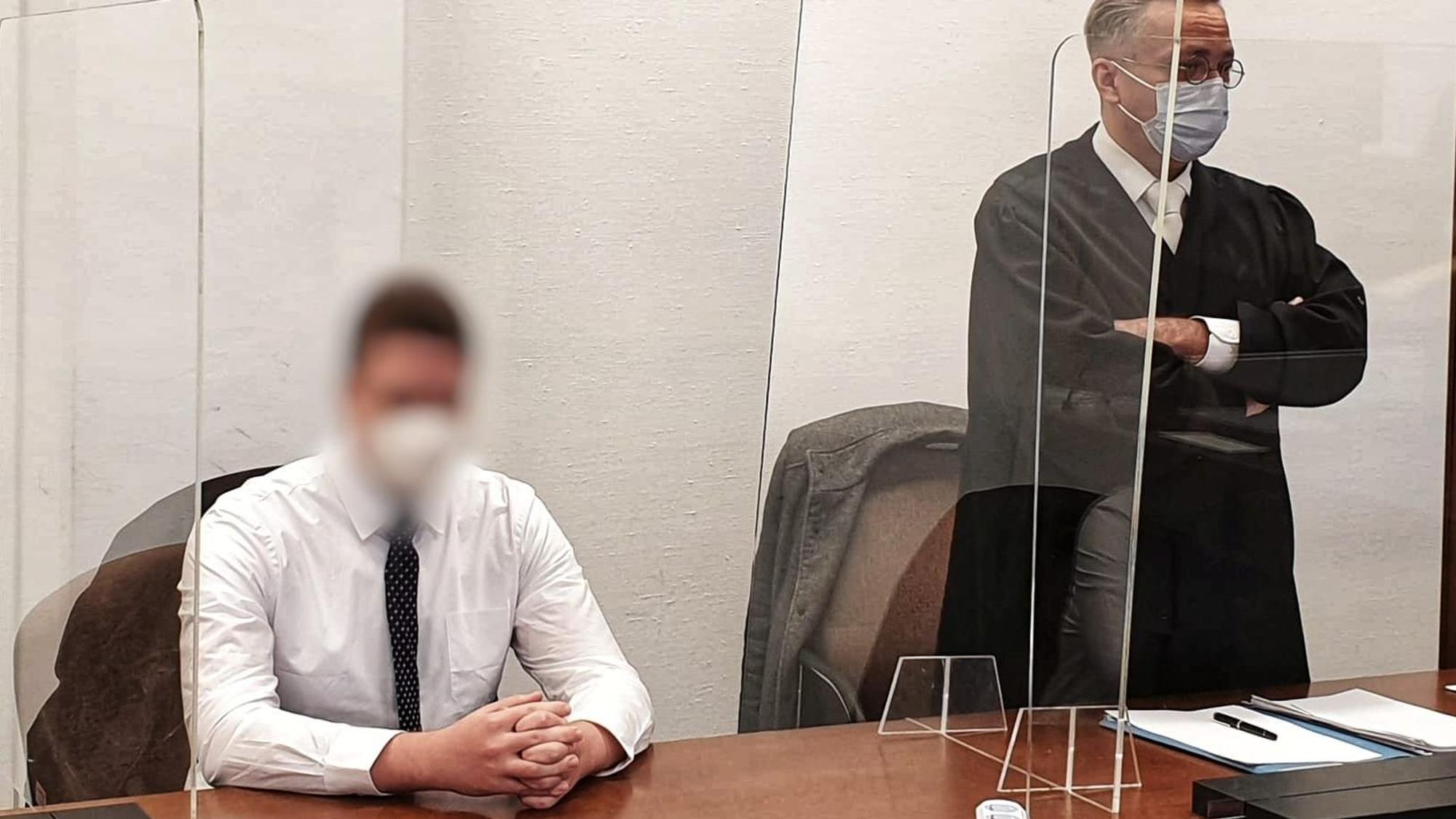 Ein junger Mann sitzt mit gefalteten Händen auf der Anklagebank, neben ihm steht sein Verteidiger.
