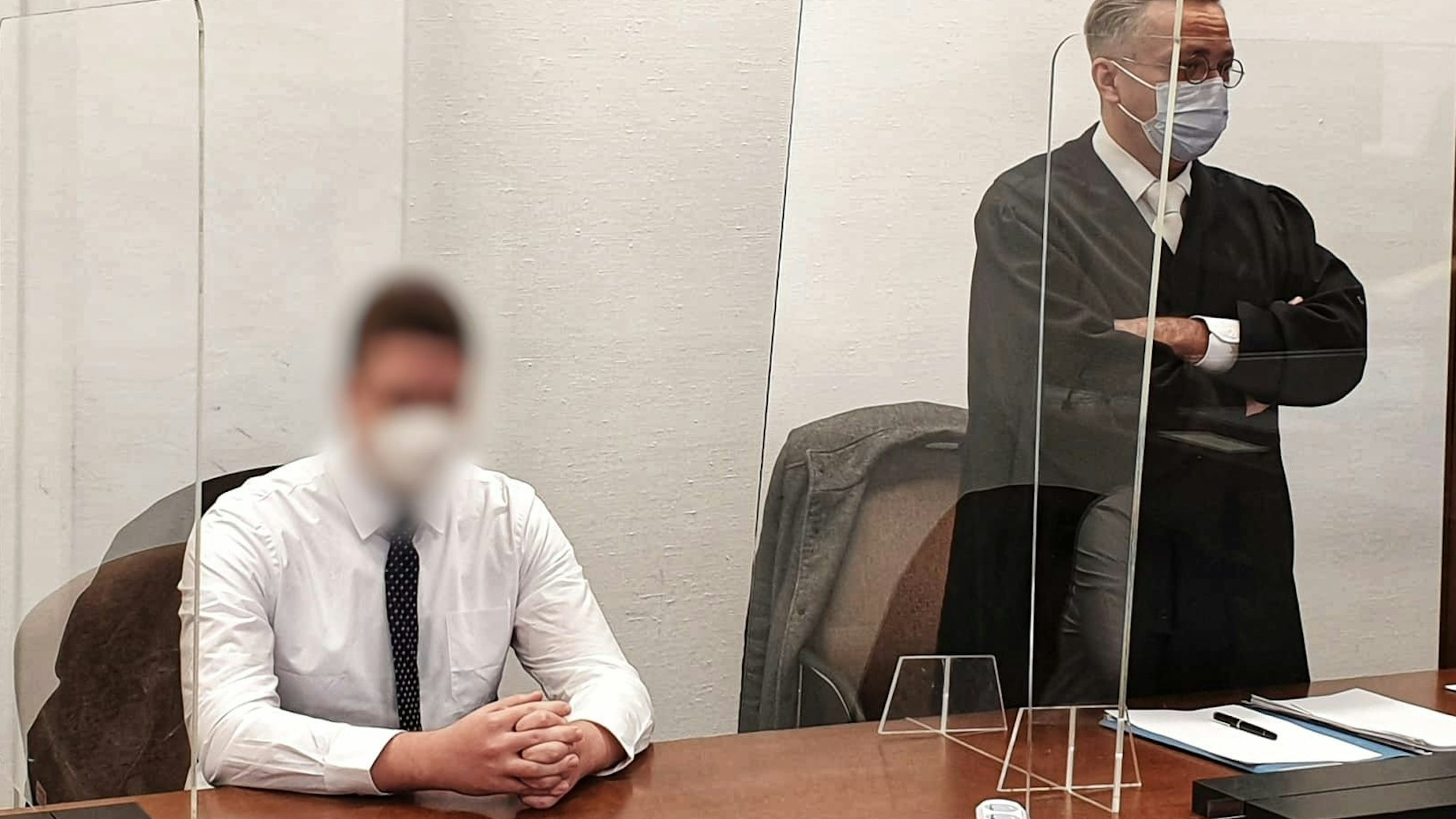 Ein junger Mann sitzt mit gefalteten Händen auf der Anklagebank, neben ihm steht sein Verteidiger.