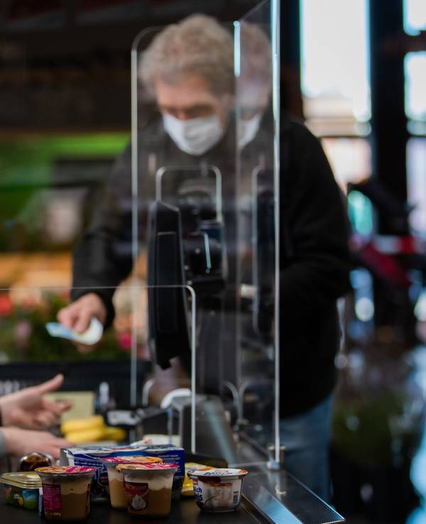 Ein Kunde mit Mundschutz bezahlt an der Kasse seinen Einkauf. An einer Kasse in einem Supermarkt wird die Kassiererin mit einer Plexiglasscheibe, einem sogenannten "Spuckschutz", geschützt.