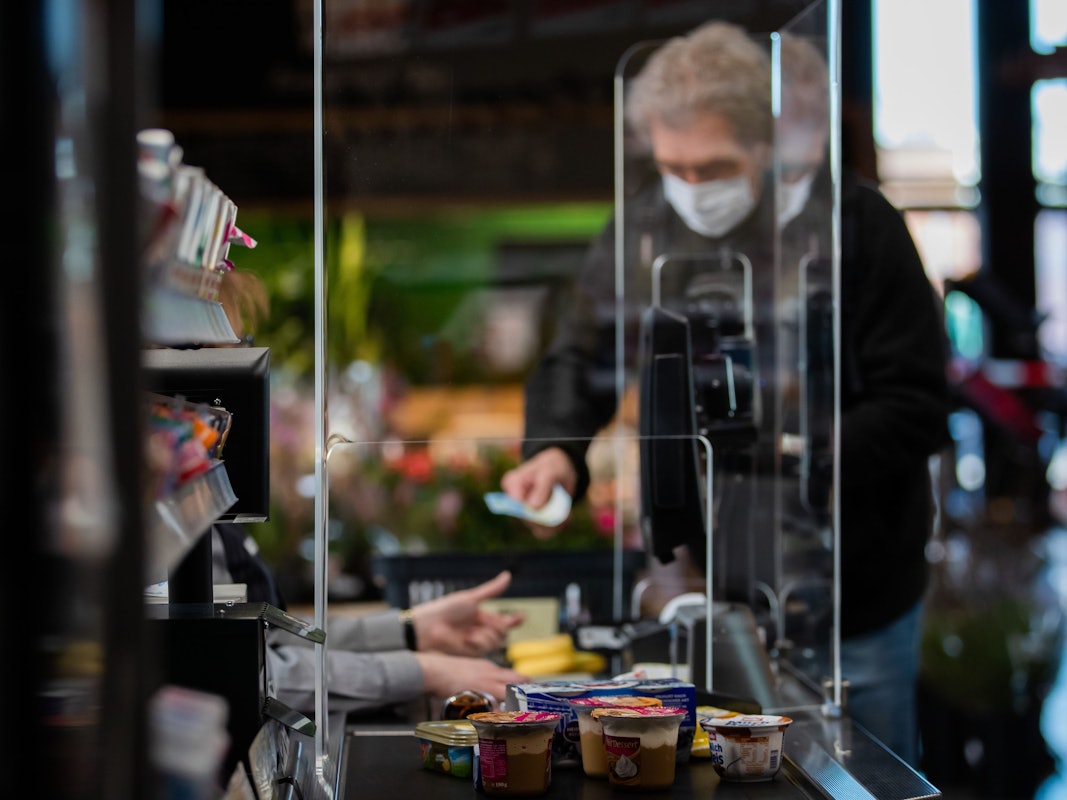 Ein Kunde mit Mundschutz bezahlt an der Kasse seinen Einkauf. An einer Kasse in einem Supermarkt wird die Kassiererin mit einer Plexiglasscheibe, einem sogenannten "Spuckschutz", geschützt.
