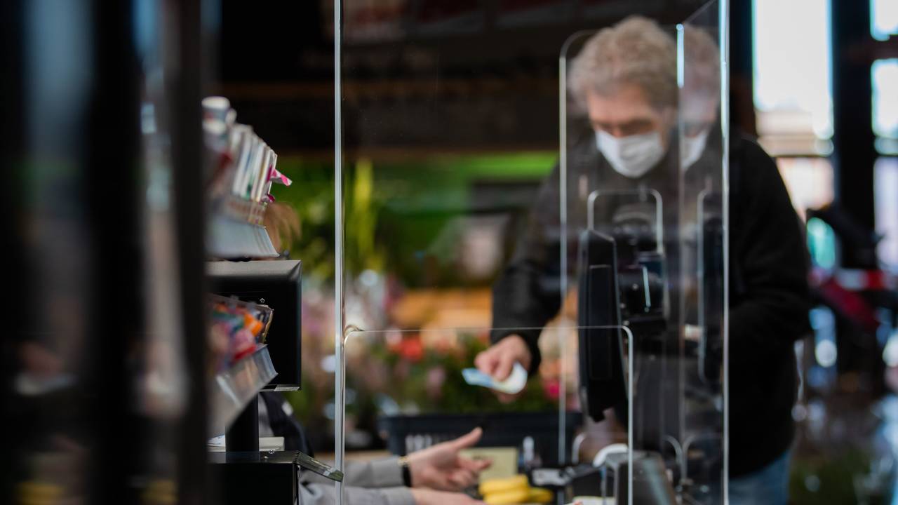 Ein Kunde mit Mundschutz bezahlt an der Kasse seinen Einkauf. An einer Kasse in einem Supermarkt wird die Kassiererin mit einer Plexiglasscheibe, einem sogenannten "Spuckschutz", geschützt.