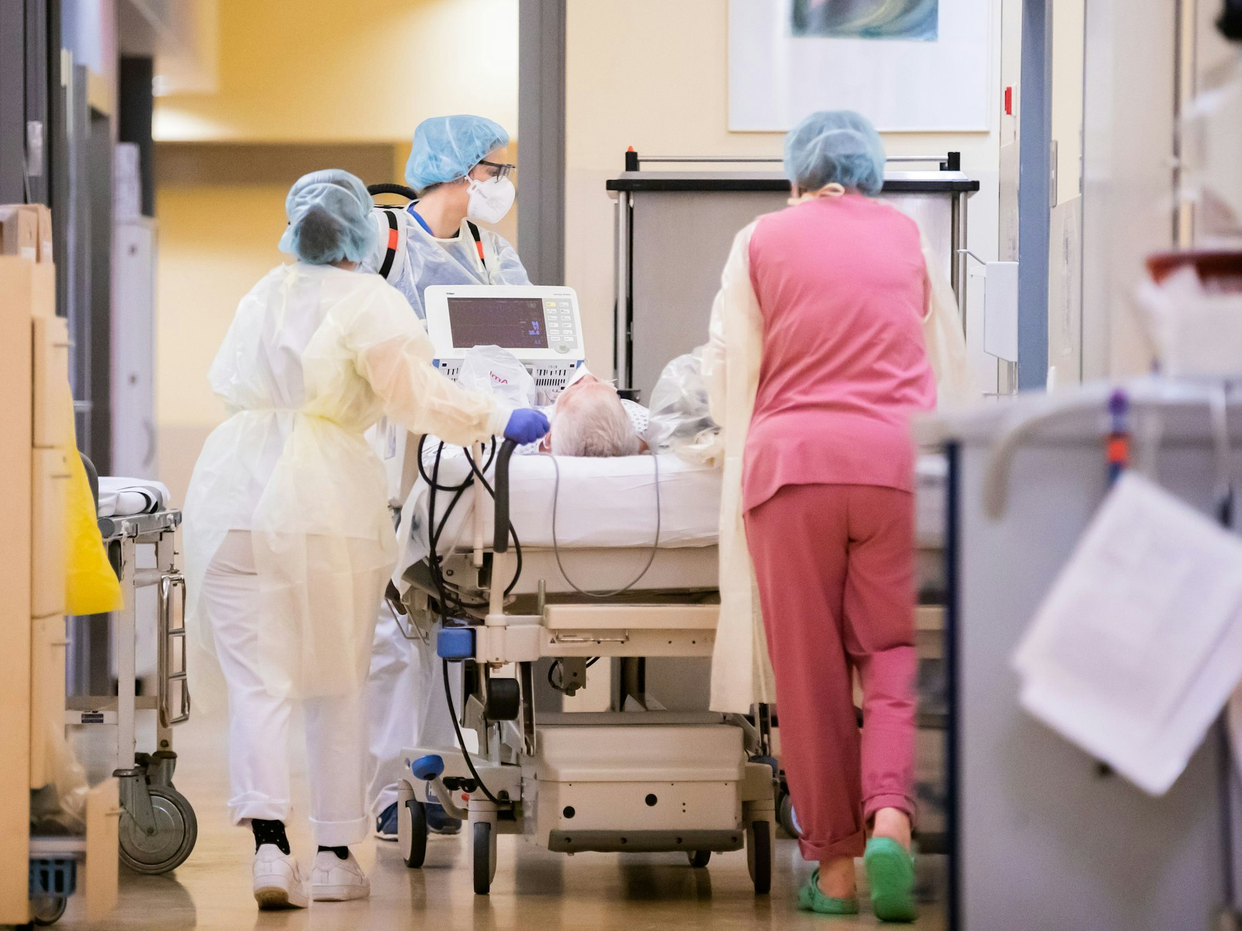 Das Symbolfoto von 2021 zeigt drei Pflegerinnen, die einen Patienten in einem Bett liegend über einen Krankenhausflur schieben.