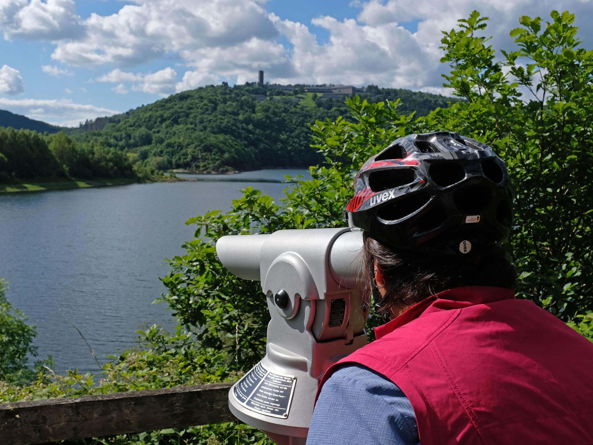 Das Foto vom 16. Juni 2019 zeigt eine Frau mit Fahrradhelm, die am Urftsee im Nationalpark Eifel durch ein Fernrohr blickt.