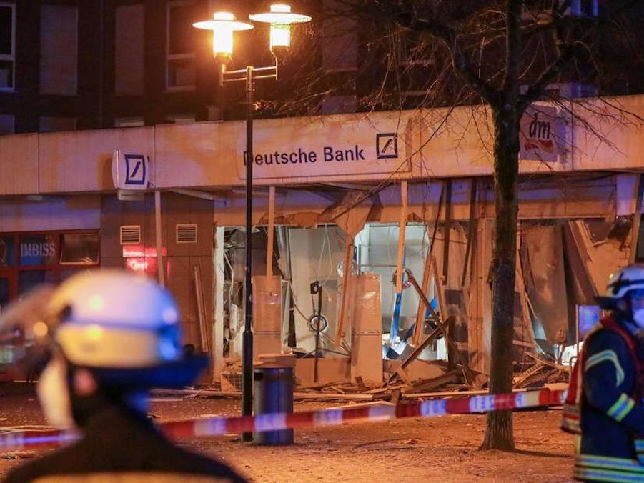 Einsatzkräfte der Feuerwehr stehen vor einer durch eine Explosion stark beschädigten Deutsche Bank-Filiale.