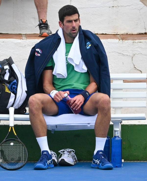 Novak Djokovic sitzt mit Jacke und Handtuch auf einer Bank, neben ihm sein Tennisschläger.