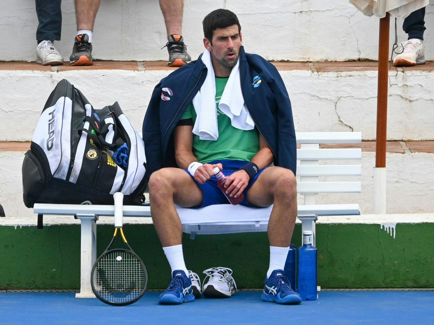 Novak Djokovic sitzt mit Jacke und Handtuch auf einer Bank, neben ihm sein Tennisschläger.