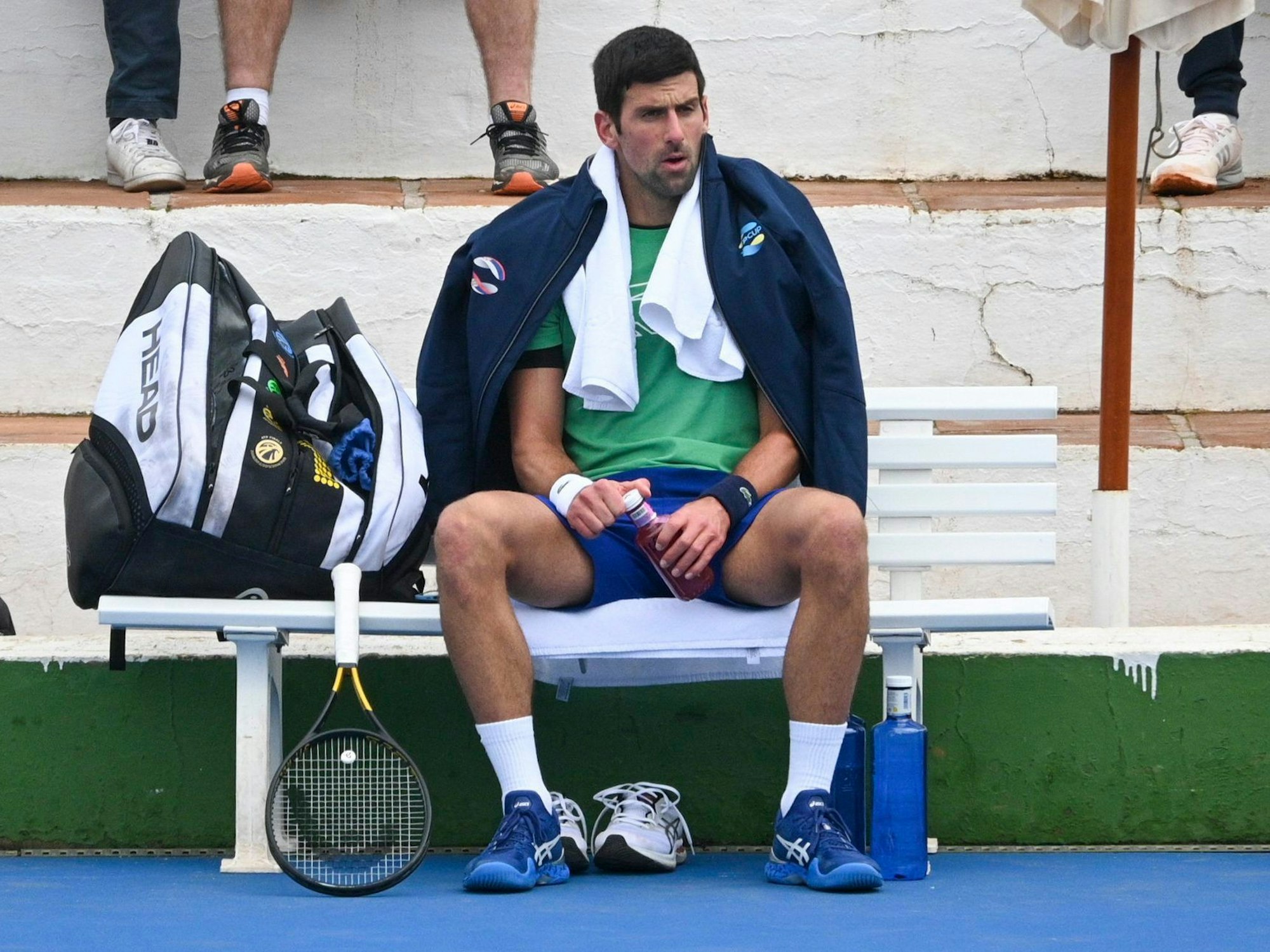 Novak Djokovic sitzt mit Jacke und Handtuch auf einer Bank, neben ihm sein Tennisschläger.