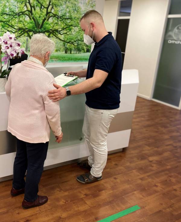Mitarbeiter einer Kölner Arztpraxis steht vor der Anmeldung und klärt eine Patientin auf.
