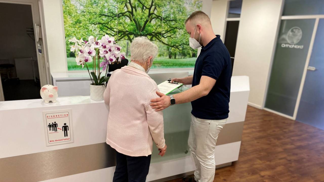 Mitarbeiter einer Kölner Arztpraxis steht vor der Anmeldung und klärt eine Patientin auf.