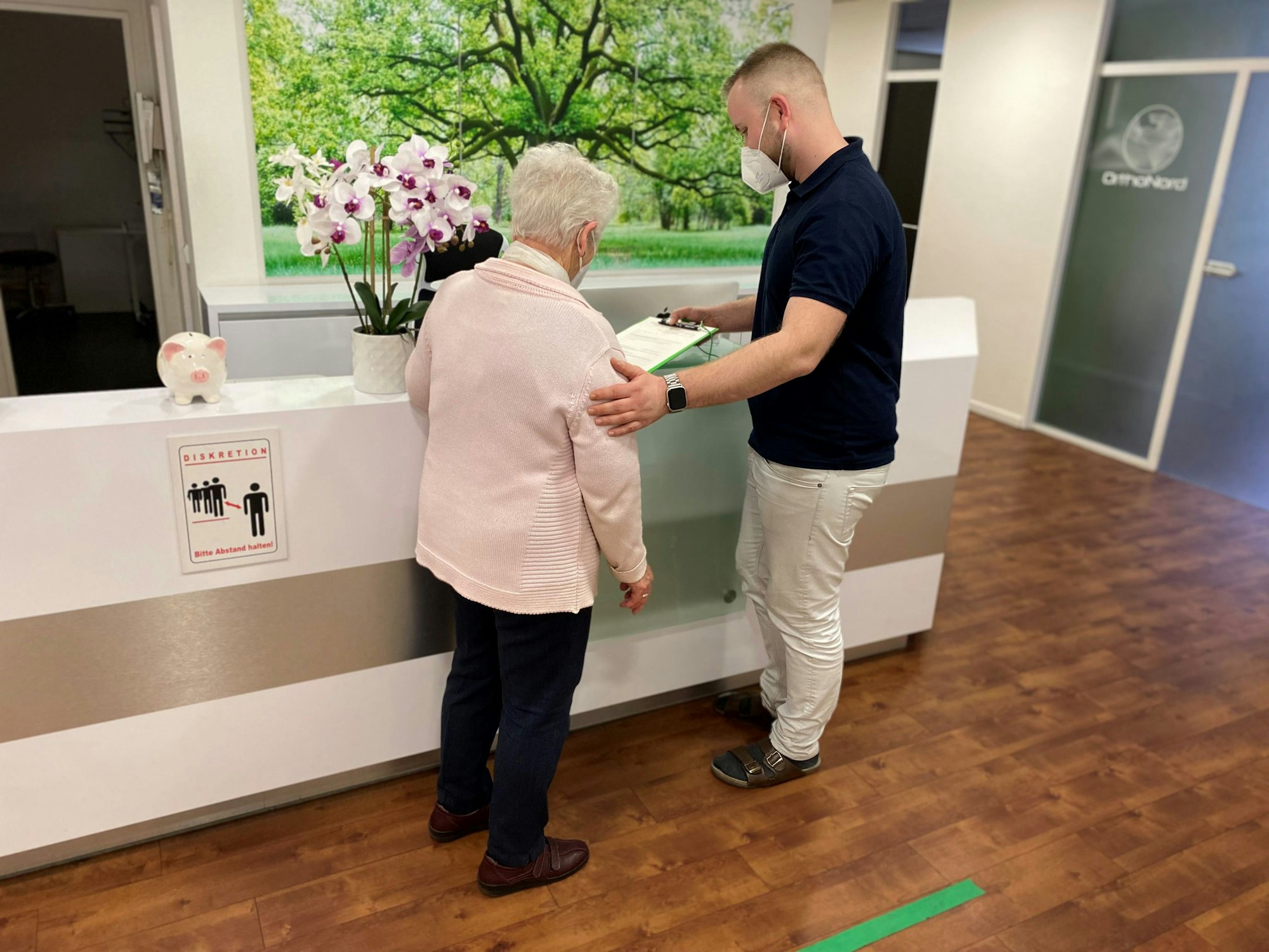 Mitarbeiter einer Kölner Arztpraxis steht vor der Anmeldung und klärt eine Patientin auf.