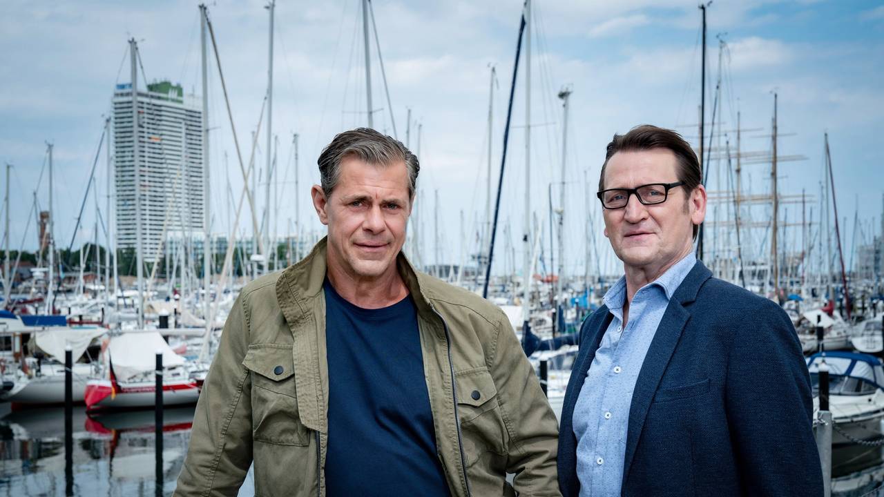 Sven Martinek (l.) und Ingo Naujoks stehen vor einem Hafen.