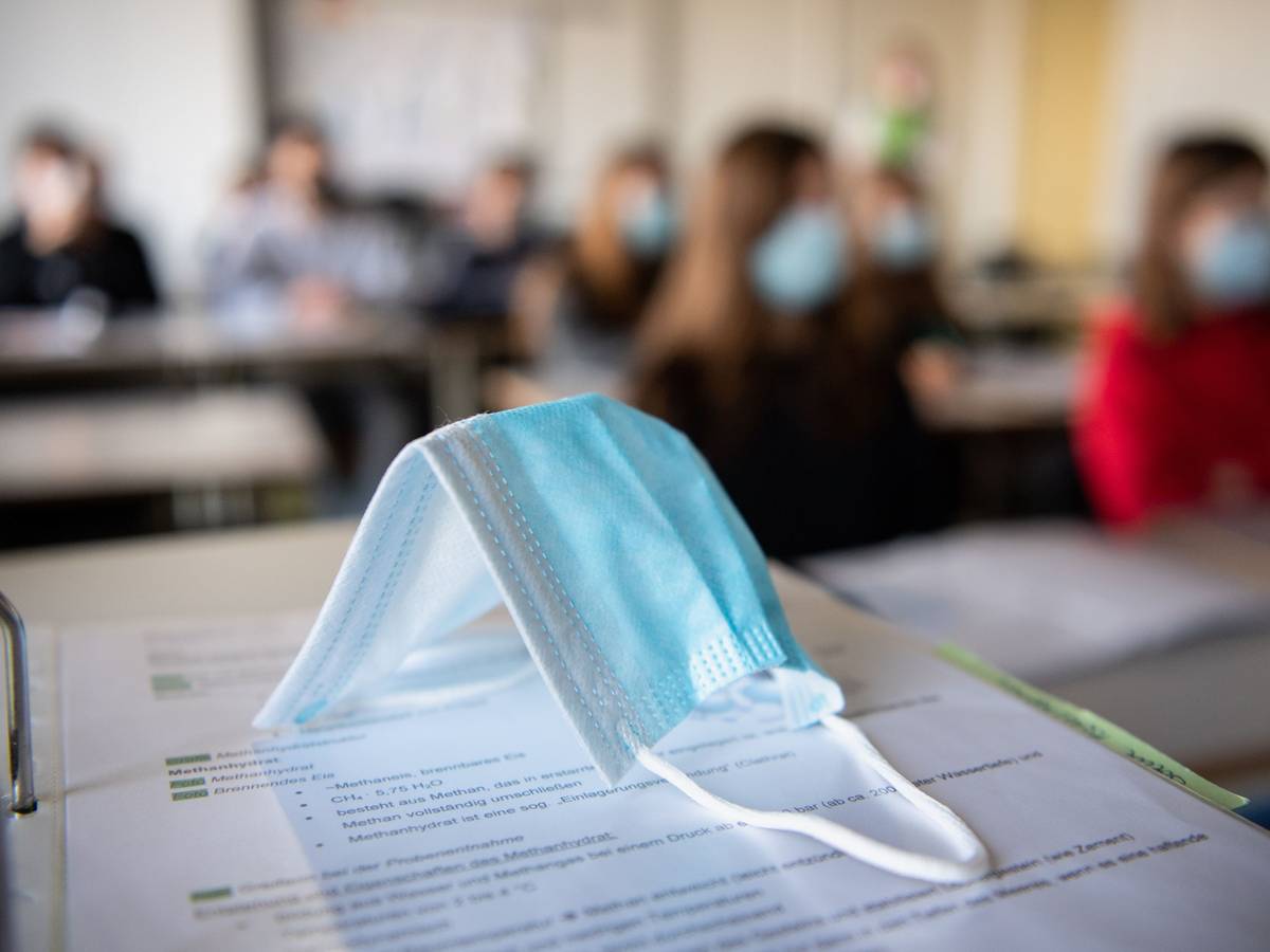 Eine Maske liegt im Unterricht auf Unterlagen, während im Hintergrund Schülerinnen und Schüler mit Mund- und Nasenschutz zu sehen sind.