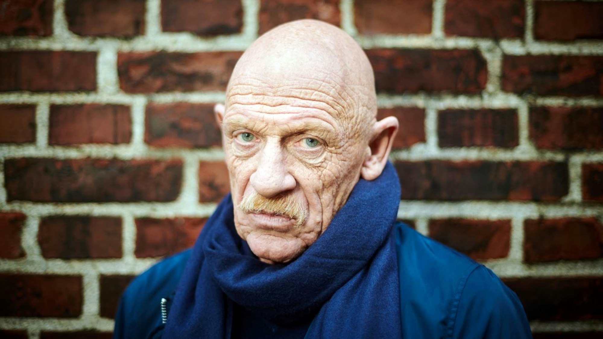 Schauspieler Joe Bausch steht vor einer Mauer und schaut in die Kamera.
