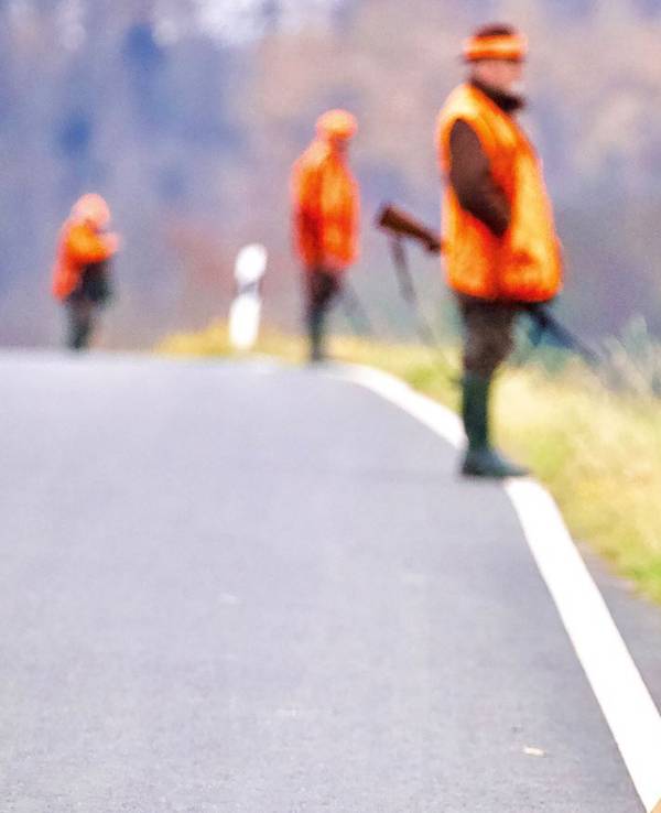 Ein Warnhinweisschild steht während einer Treibjagd am Rande einer Straße. Im Hintergrund stehen Jäger mit Gewehren.