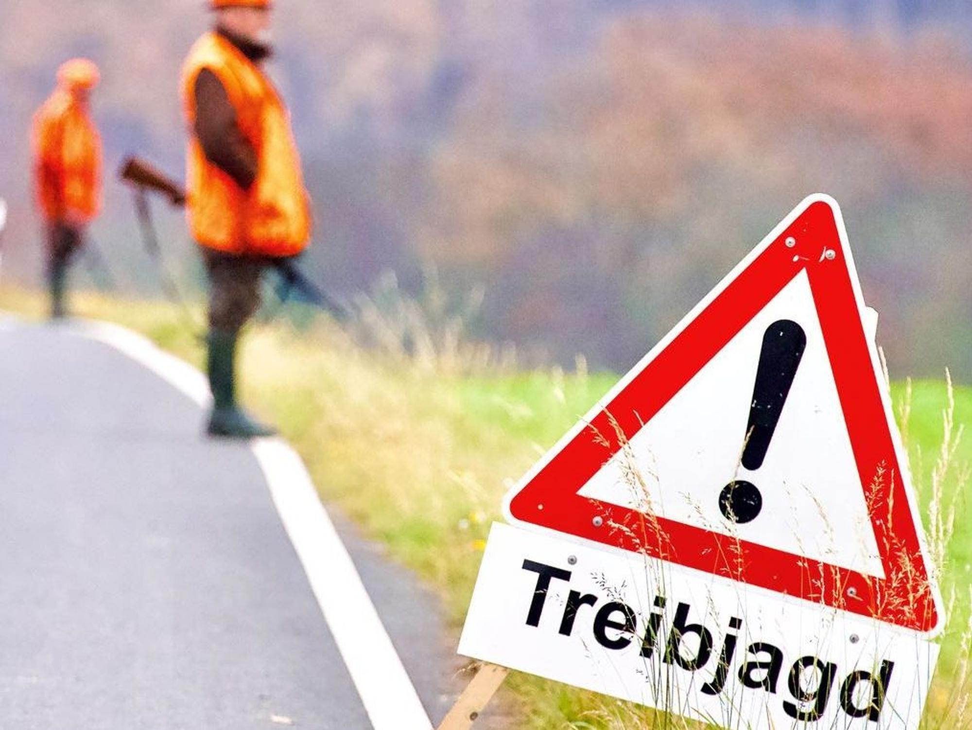 Ein Warnhinweisschild steht während einer Treibjagd am Rande einer Straße. Im Hintergrund stehen Jäger mit Gewehren.