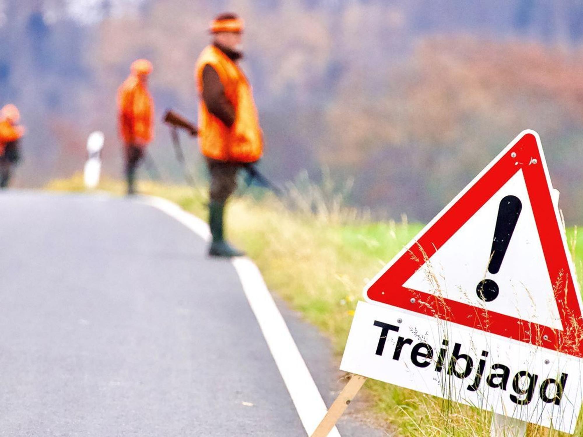 Ein Warnhinweisschild steht während einer Treibjagd am Rande einer Straße. Im Hintergrund stehen Jäger mit Gewehren.
